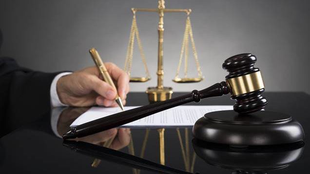 Close-up Of Judge Writing On Paper With Gavel And Scale At Desk